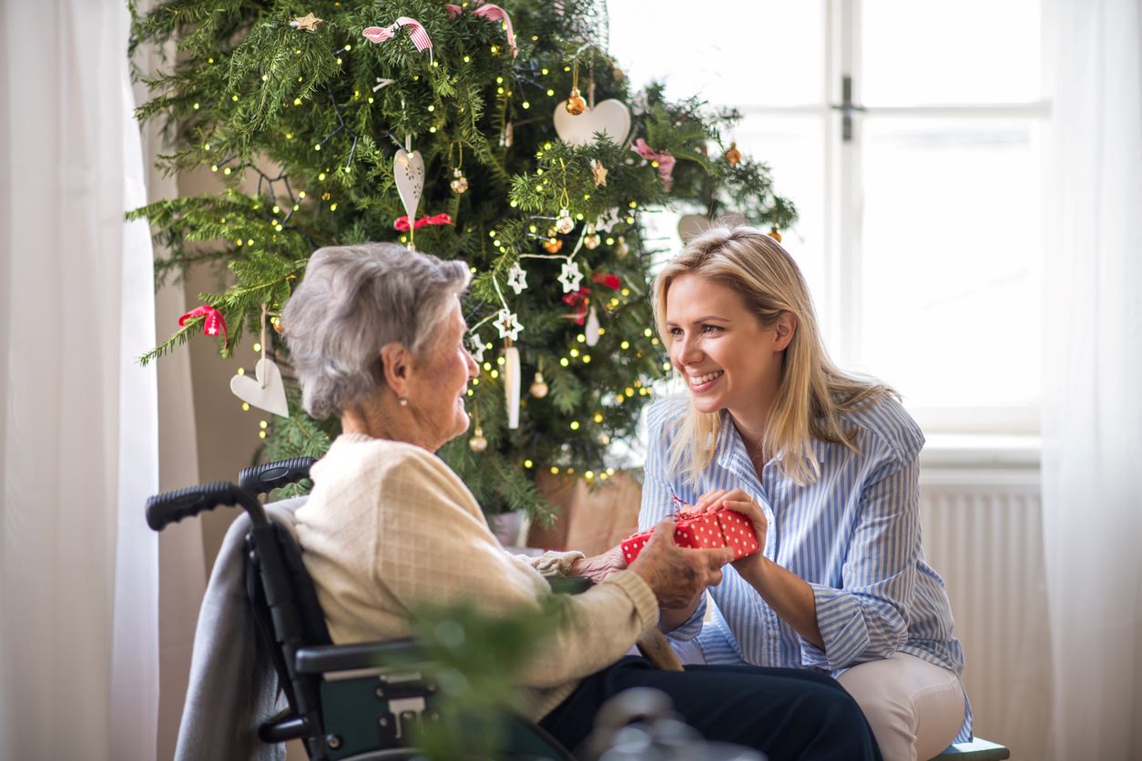 A smiling young woman and an older woman in a wheelchair exchange a gift in front of a Christmas tree