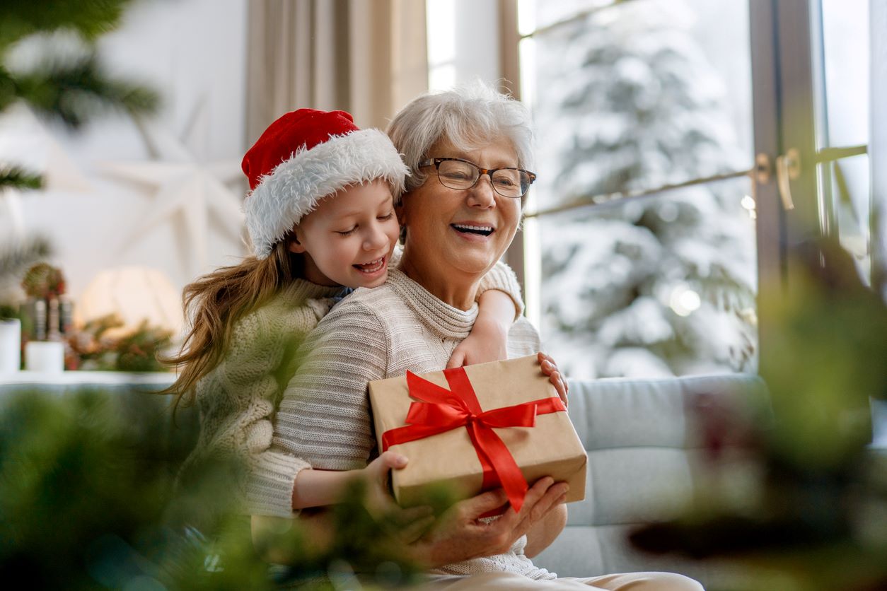 A smiling grandchild in a Santa hat hugs her grandmother from behind while giving a Christmas gift.