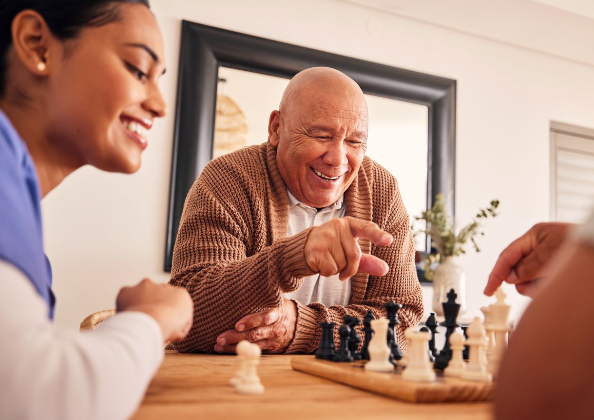 A senior man enjoys playing chess as a caregiver smiles and watches.