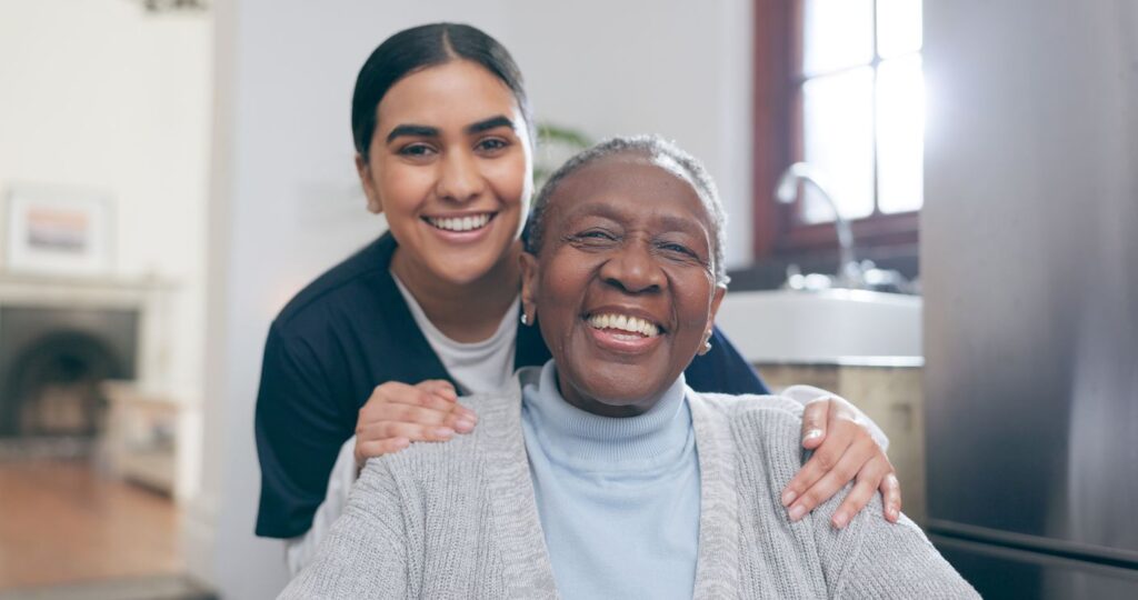 A senior woman sits with her caregiver standing behind her, both smiling.