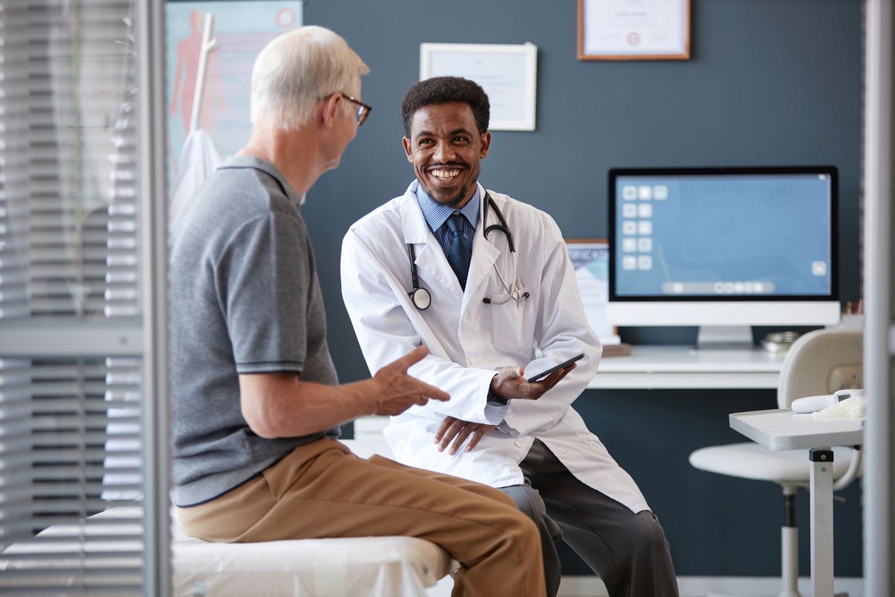 A smiling doctor holds a tablet while talking to a patient in an exam room with a computer on a desk behind