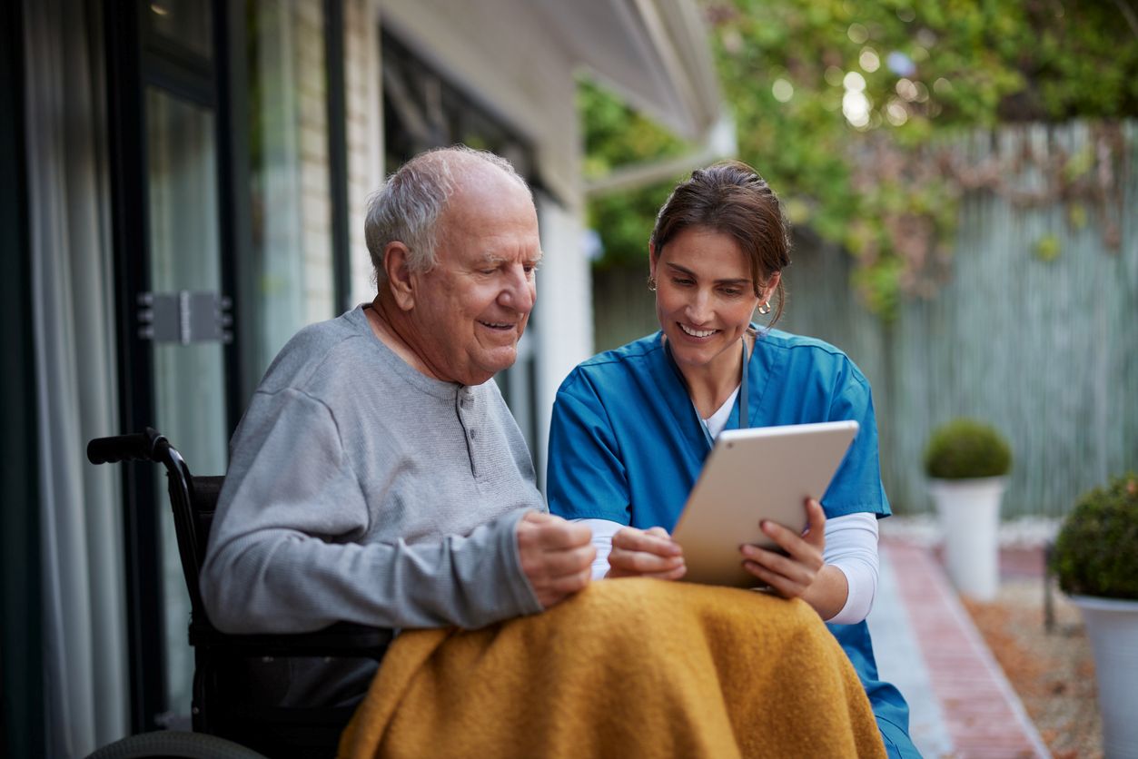 A man in a wheelchair receives assistance with an iPad from an in-home caregiver.