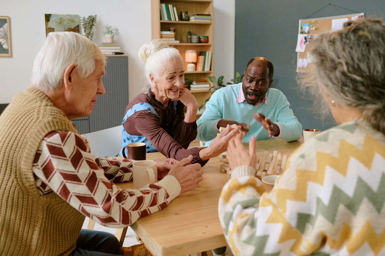 A group of seniors drink coffee and play Jenga around a wooden table.
