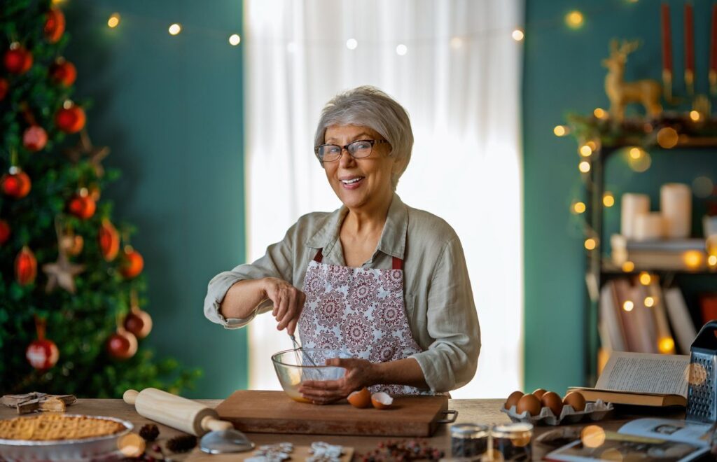 A smiling senior woman mixes cookie batter on the counter in front of her Christmas tree
